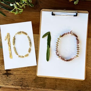Pode incluir: Dois cart&otilde;es brancos e uma prancheta com o n&uacute;mero 10. Um cart&atilde;o tem o n&uacute;mero 10 feito de flores e caules secos. O outro cart&atilde;o tem o n&uacute;mero 10 feito de contas e caules dourados. A prancheta tem o n&uacute;mero 10 feito de folhas e flores.