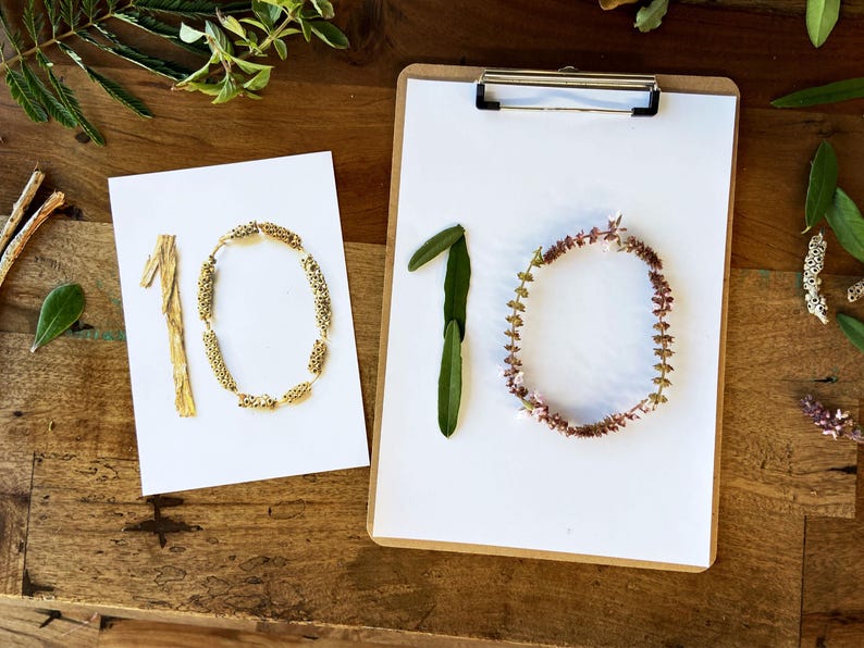 Pode incluir: Dois cart&otilde;es brancos e uma prancheta com o n&uacute;mero 10. Um cart&atilde;o tem o n&uacute;mero 10 feito de flores e caules secos. O outro cart&atilde;o tem o n&uacute;mero 10 feito de contas e caules dourados. A prancheta tem o n&uacute;mero 10 feito de folhas e flores.
