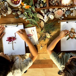 May include: Two children are creating art with natural materials. One child is making a flamingo out of pink flowers and twigs, while the other is making a turtle out of leaves and stones.