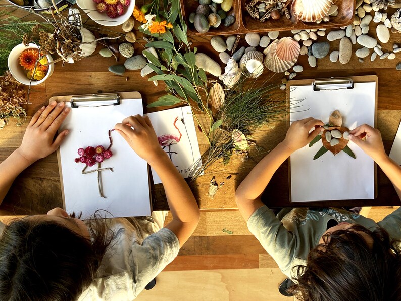 May include: Two children are creating art with natural materials. One child is making a flamingo out of pink flowers and twigs, while the other is making a turtle out of leaves and stones.