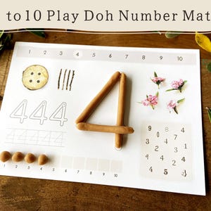 Könnte beinhalten: Eine weiße Spielunterlage mit den Zahlen 1 bis 10. Die Zahl vier ist mit brauner Knete geformt. Die Unterlage enthält Illustrationen einer Knopfs, Linien und Blumen.