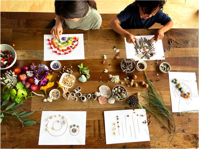 May include: Two children create nature art on white paper. One child uses colorful flower petals to make a rainbow shape. The other child uses twigs and other natural materials to create a bird's nest. The table is covered in a variety of natural materials, including flowers, shells, and stones.