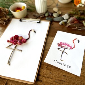 May include: Two paper cards with pink and red flower arrangements depicting flamingos. The cards are on a wooden surface. The card on the left has the text "flamingo" written on it.