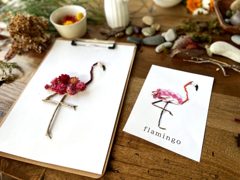 May include: Two paper cards with pink and red flower arrangements depicting flamingos. The cards are on a wooden surface. The card on the left has the text "flamingo" written on it.