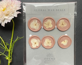 May include: A sheet of self-adhesive floral wax seals with dried flowers pressed into the wax. The seals are a light pink color and feature white flowers. The text "SELF-ADHESIVE FLORAL WAX SEALS" is printed on the sheet. The text "FOUND FLORAL CO" is printed at the bottom of the sheet.