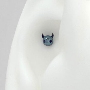 May include: A small, gray, devil-shaped stud earring with black horns and eyes. The earring is shown in a close-up image on a white ear model.