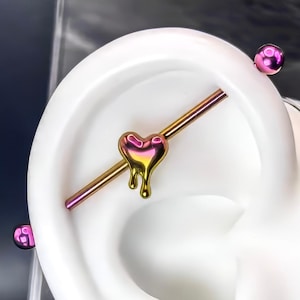 May include: A gold heart-shaped barbell with a pink and purple iridescent finish. The barbell is inserted into a person's earlobe.