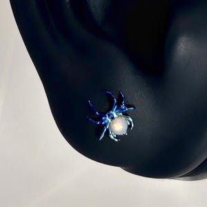 May include: A blue spider stud earring with a white center is displayed on a black ear-shaped display. The earring has a detailed spider design. The text "- Ti Candy Un -" is visible in the upper left corner.