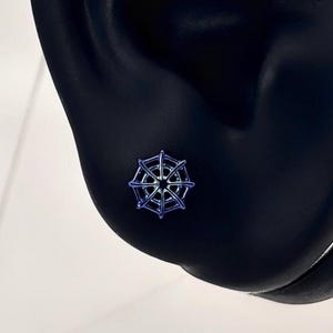 May include: A small, blue and silver stud earring in the shape of a ship's wheel is displayed on a black ear model. The earring has a detailed design with a dark blue center and silver spokes. The ear model is set against a white background.