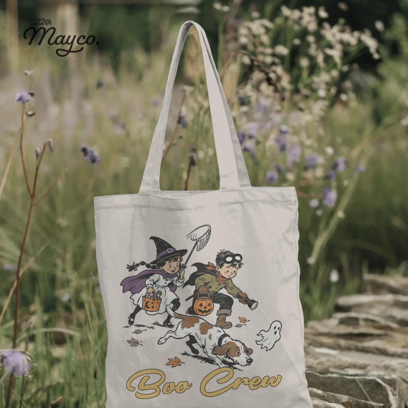 May include: A cream-colored canvas tote bag featuring a vintage-style illustration of a child dressed as a witch and another as an explorer with a dog, a small ghost, and the text "Boo Crew" printed in yellow. The brand "22th Mayco." is visible.