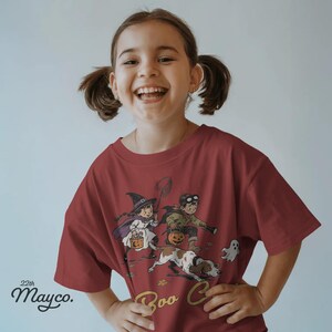 May include: A dark red short-sleeved t-shirt featuring a graphic illustration of two children in Halloween costumes, a dog, and a ghost. The text 22th Mayco. is printed in the bottom left corner. The graphic includes a witch and a child with goggles.