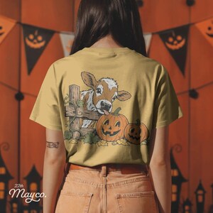 May include: A mustard yellow t-shirt featuring a graphic print on the back. The illustration depicts a brown and white calf peeking over a wooden fence next to two orange jack-o'-lantern pumpkins with carved faces and green leaves.
