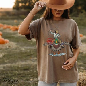 May include: Brown t-shirt with a graphic of a cowboy riding a duck, holding a lasso. The duck has outstretched wings and is standing in water. The text "22nd Mayco" is at the bottom left.