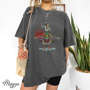 May include: Dark gray t-shirt featuring a graphic of a cowboy riding a duck, with the text "DO NOT COPY 22TH MAYCO" in a circular design. The shirt has a vintage, washed look.