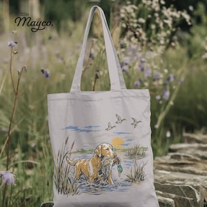 May include: A light-colored canvas tote bag featuring a graphic illustration of a golden retriever standing in water with a duck in its mouth. The scene includes cattails, a yellow sun, and three flying ducks. The text 22th Mayco. is printed in black.