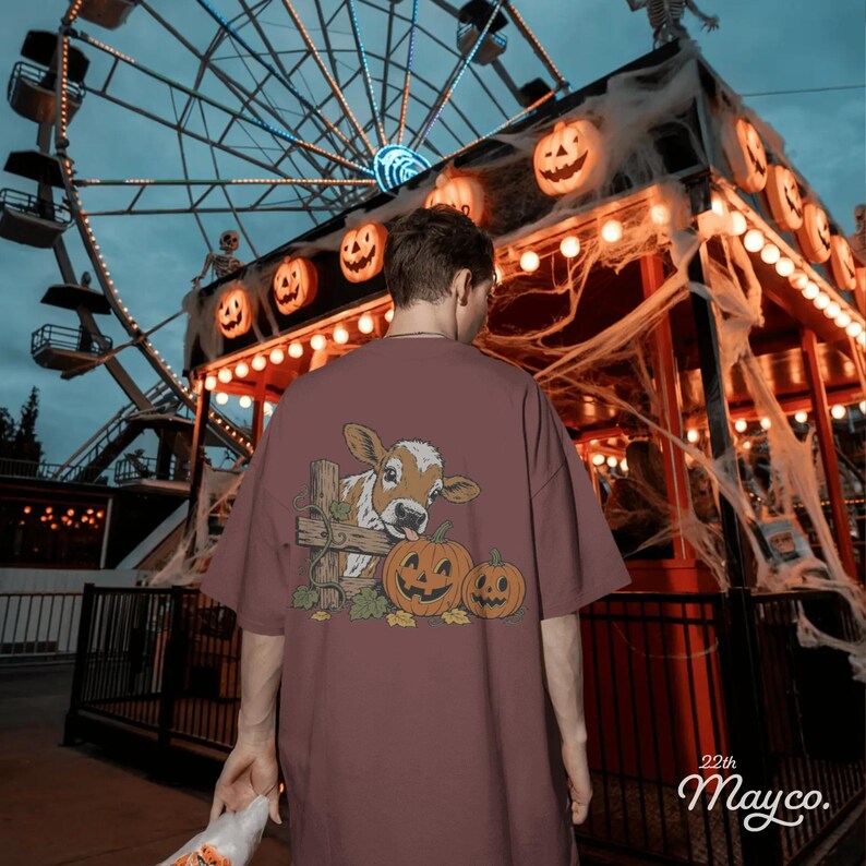 May include: A dusty rose short-sleeve t-shirt featuring a graphic on the back of a brown and white calf peeking over a wooden fence next to two jack-o'-lantern pumpkins with carved faces, surrounded by green leaves and vines.