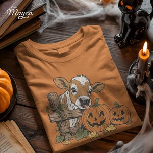 May include: A mustard yellow t-shirt featuring a graphic illustration of a brown and white calf peeking over a wooden fence next to two orange jack-o'-lantern pumpkins with carved faces, green leaves, and vines, printed on the front of the shirt.