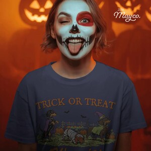 May include: A dark blue t-shirt featuring a graphic of three dachshunds in Halloween costumes with the text TRICK OR TREAT. One dog wears a purple cape, one is wrapped like a mummy, and one wears a green cape and a witch hat in a pumpkin patch.