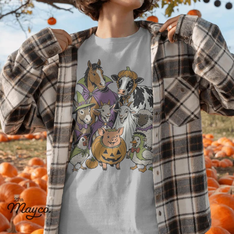 K&ouml;nnte beinhalten: Ein hellgraues T-Shirt mit einer grafischen Illustration von Bauernhoftieren in Halloween-Kost&uuml;men, darunter ein Pferd im Umhang, eine Kuh mit K&uuml;rbishut, ein Schwein im K&uuml;rbiskost&uuml;m und eine Ente als Frankensteins Monster.