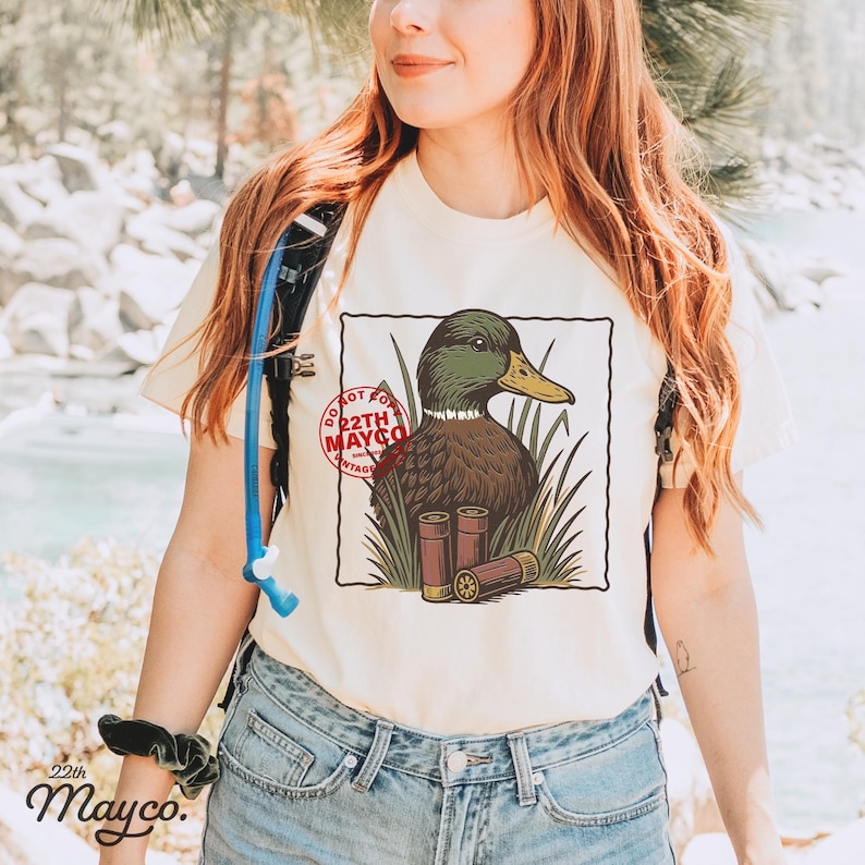 K&ouml;nnte beinhalten: Cremefarbenes T-Shirt mit einer detaillierten Illustration einer Ente mit gr&uuml;nen und braunen Federn, einem gelben Schnabel und zwei Schrotpatronen. Das Design enth&auml;lt den Text "DO NOT FOR 22TH MAYCO VINTAGE". Das Shirt wird mit einer blauen Jeans kombiniert.
