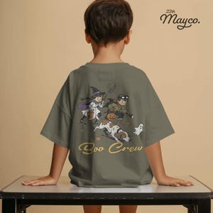 May include: An olive green t-shirt featuring a graphic print on the back. The illustration depicts two children in Halloween costumes, one as a witch and one as an explorer, with a dog and a small ghost. The text "Boo Crew" is printed below.