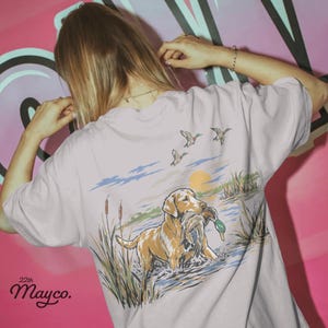 May include: A light lavender t-shirt featuring a graphic on the back of a golden retriever standing in water holding a duck in its mouth. The scene includes cattails, a setting sun, and three flying ducks. The text 22th Mayco. is printed in black.