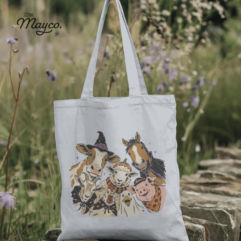 May include: A white canvas tote bag featuring a graphic illustration of a cow wearing a witch hat, a horse, a sheep, a goat, a pig, and a duck. The text 22th Mayco. is printed in the upper left corner above the farm animals in black ink.