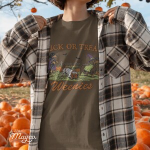 May include: A brown t-shirt featuring a graphic of three dachshunds in Halloween costumes with pumpkins and bats. The text reads TRICK OR TREAT Weenies. The shirt is worn under a brown, white, and black plaid button-down long-sleeve flannel shirt.