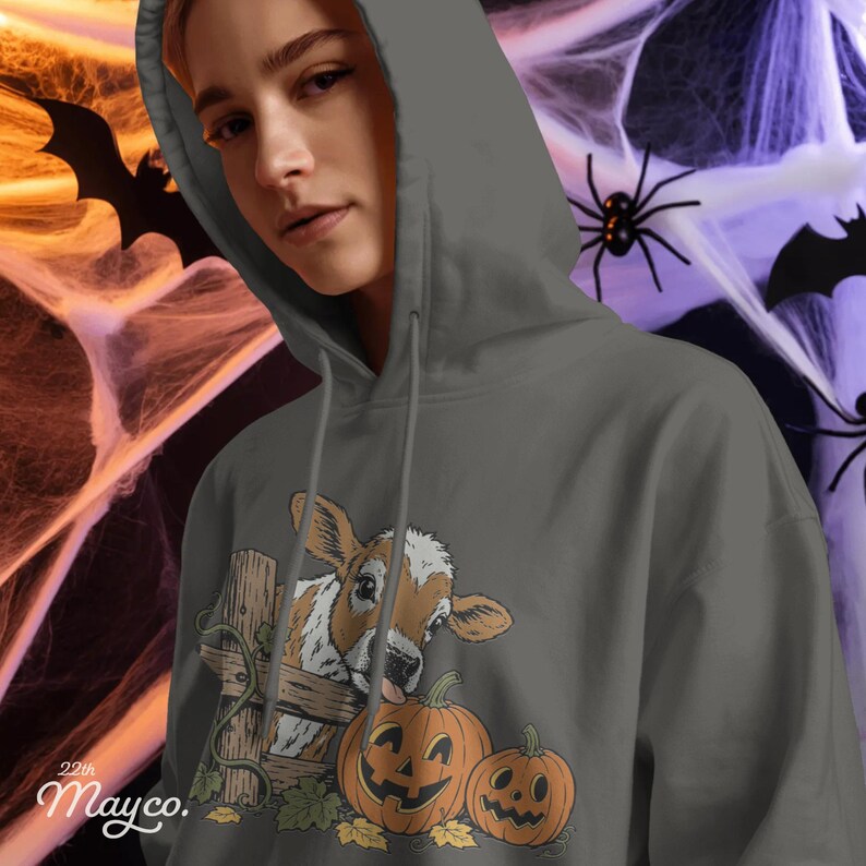 May include: A dark gray hooded sweatshirt featuring a graphic print of a brown and white calf peeking over a wooden fence next to two orange jack-o'-lantern pumpkins. The text "22th Mayco." is printed in white in the bottom left corner.