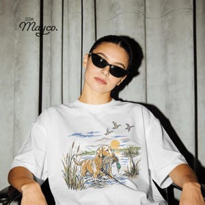 May include: A white t-shirt featuring a graphic illustration of a golden retriever standing in water with a duck in its mouth, surrounded by reeds and flying ducks under a yellow sun. The text 22th Mayco. is printed in black in the upper left corner.