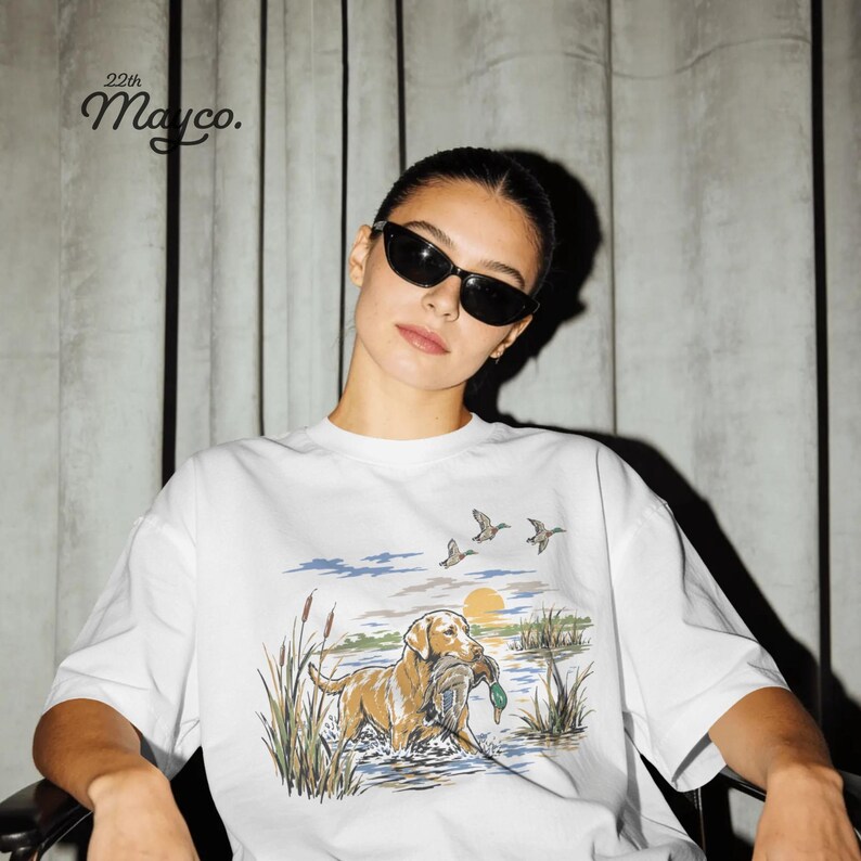 May include: A white t-shirt featuring a graphic illustration of a golden retriever standing in water with a duck in its mouth, surrounded by reeds and flying ducks under a yellow sun. The text 22th Mayco. is printed in black in the upper left corner.