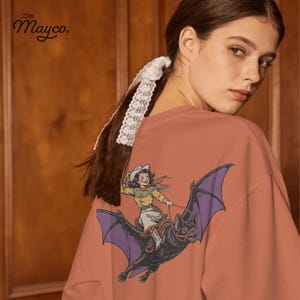 vintage halloween cowgirl png, retro western rider illustration, hand drawn giant bat graphic, purple orange yellow halloween palette, smiling cowgirl with cowboy hat, fringed western shirt and orange scarf, flying bat saddle and reins design, classic cowgirl adventure graphic, spooky rodeo halloween png, bold outline sublimation design, nostalgic western girl illustration, detailed purple bat wings, country fall shirt graphic, transparent png digital download, no distressed halloween design