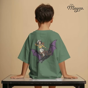 May include: A sage green short-sleeve t-shirt featuring a graphic on the back of a person in western attire riding a large black bat with purple wings. The text 22th Mayco. is printed in the upper right corner of the shirt in black lettering.
