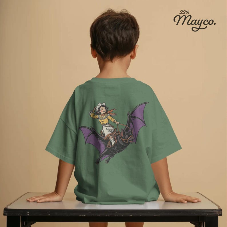 May include: A sage green short-sleeve t-shirt featuring a graphic on the back of a person in western attire riding a large black bat with purple wings. The text 22th Mayco. is printed in the upper right corner of the shirt in black lettering.