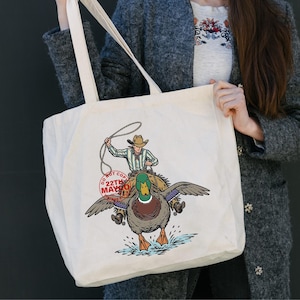 May include: Cream-colored tote bag featuring a cowboy riding a duck, with a lasso in hand. The duck has outstretched wings and is splashing in water. The text "DO NOT COPY 22TH MAY" is printed on a red stamp.