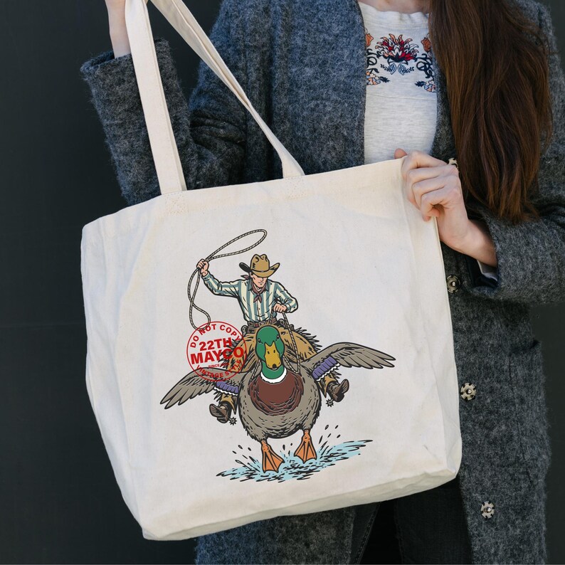 May include: Cream-colored tote bag featuring a cowboy riding a duck, with a lasso in hand. The duck has outstretched wings and is splashing in water. The text "DO NOT COPY 22TH MAY" is printed on a red stamp.