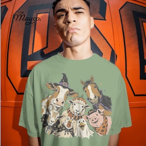 May include: A sage green t-shirt featuring a graphic illustration of farm animals dressed in Halloween costumes, including a cow in a witch hat, a horse in a cape, a sheep in a pumpkin hat, a goat, a chicken, a duck, and a pig in a bandana.