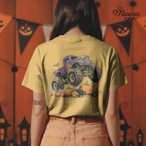 Puede incluir: Una camiseta color amarillo mostaza con un gr&aacute;fico de un cami&oacute;n monstruo morado con neum&aacute;ticos grandes pasando sobre calabazas naranjas. El cami&oacute;n tiene un peque&ntilde;o icono de calabaza en la puerta. El texto 22th Mayco. aparece en la esquina superior.