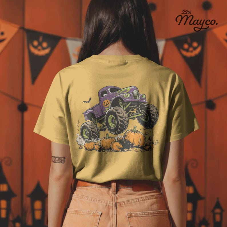 Puede incluir: Una camiseta color amarillo mostaza con un gr&aacute;fico de un cami&oacute;n monstruo morado con neum&aacute;ticos grandes pasando sobre calabazas naranjas. El cami&oacute;n tiene un peque&ntilde;o icono de calabaza en la puerta. El texto 22th Mayco. aparece en la esquina superior.