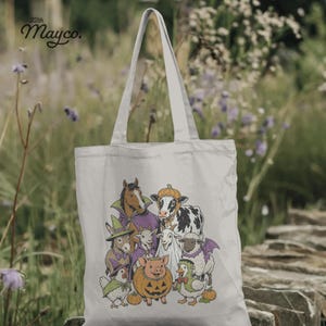 K&ouml;nnte beinhalten: Eine helle Stofftasche mit einer grafischen Illustration von Bauernhoftieren in Halloween-Kost&uuml;men, darunter ein Pferd, eine Kuh, ein Schwein, ein Schaf, eine Ziege und ein Huhn. Der Text 22th Mayco. ist in der oberen linken Ecke aufgedruckt.