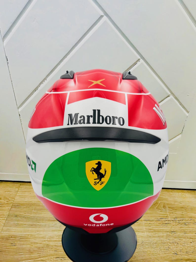 May include: A white, red, and green motorcycle helmet with a black visor. The helmet has the Marlboro logo, a Ferrari logo, and the Vodafone logo.