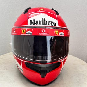 May include: Red motorcycle helmet with a black visor and a white Marlboro logo. The helmet also has smaller logos for Vodafone and Ferrari.