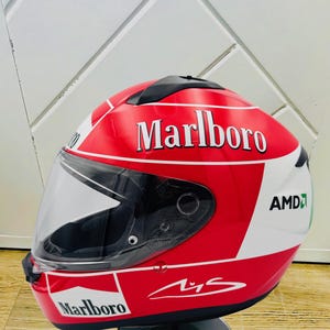 May include: A red, white, and green motorcycle helmet with a Marlboro logo and a signature. The helmet has a clear visor and is on a black stand.