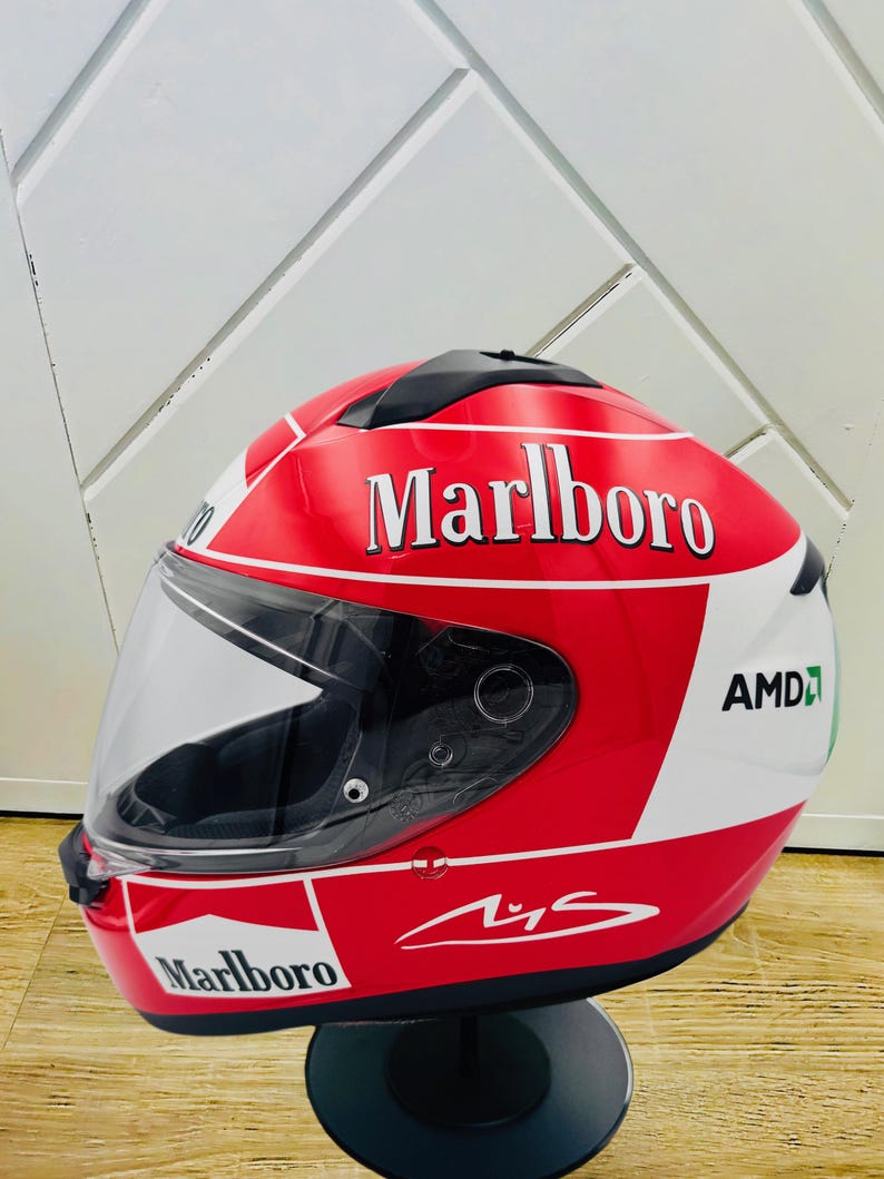 May include: A red, white, and green motorcycle helmet with a Marlboro logo and a signature. The helmet has a clear visor and is on a black stand.