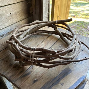 May include: A rustic, circular wreath crafted from intertwined brown branches. The wreath is tied with twine and rests on a weathered wooden surface, suggesting a natural, handcrafted aesthetic. Ideal for home decor.