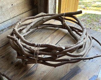 May include: A rustic, circular wreath crafted from intertwined brown branches. The wreath is tied with twine and rests on a weathered wooden surface, suggesting a natural, handcrafted aesthetic. Ideal for home decor.