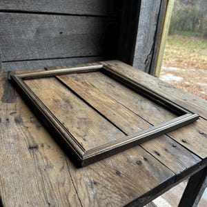 May include: A simple, rectangular picture frame made of wood with a gold finish. The frame is lying on a wooden surface.