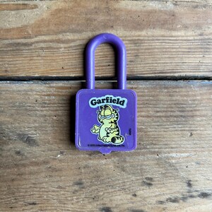May include: A purple Garfield padlock featuring the cartoon cat Garfield. The padlock has a rectangular body with the Garfield logo and a cartoon image of Garfield. The lock's handle is a matching purple color.