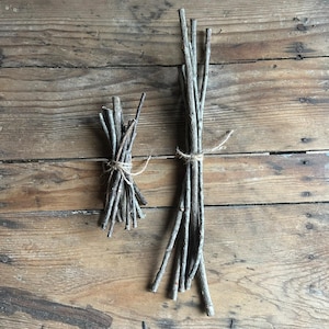 May include: Two bundles of thin, gray-brown twigs tied with twine, displayed on a weathered wooden surface. The twigs vary in length, with one bundle being smaller than the other. The natural arrangement suggests a rustic, decorative theme.