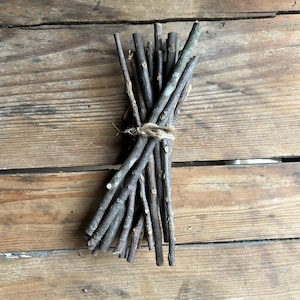 May include: A bundle of thin, dark gray twigs tied together with twine. The twigs are of varying lengths and diameters, creating a natural, rustic aesthetic. The bundle rests on a weathered wooden surface.